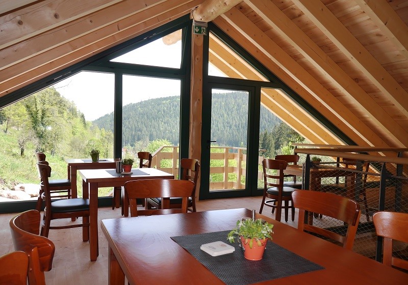 Innenraum einer Holzhütte mit großer Fensterfront davor stehen Tische und Stühle.